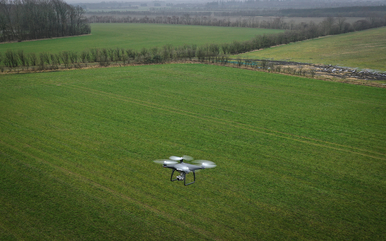 En drone med kamera flyver over grønne marker.
